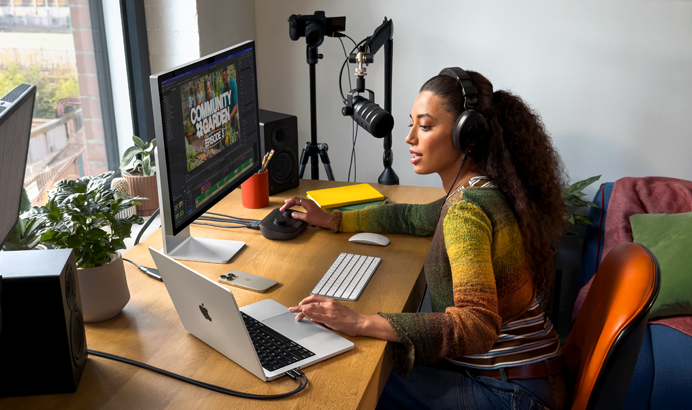 Een vrouw werkt thuis aan haar bureau met een MacBook Pro die is verbonden met twee externe displays, ze draagt oortjes en praat in een microfoon